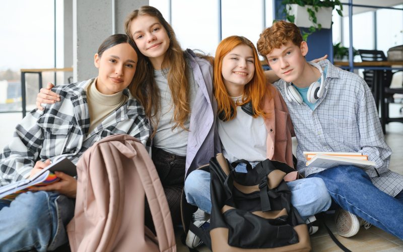 A group of cheerful student friends are sitting on the floor in a classroom.