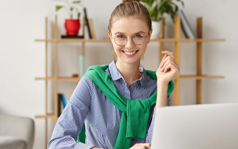 Beautiful female specialist or programmer tries to understand new computer programm and how it works, smiles pleasantly as acieves great results in sphere of innovative technologies, rejoices success