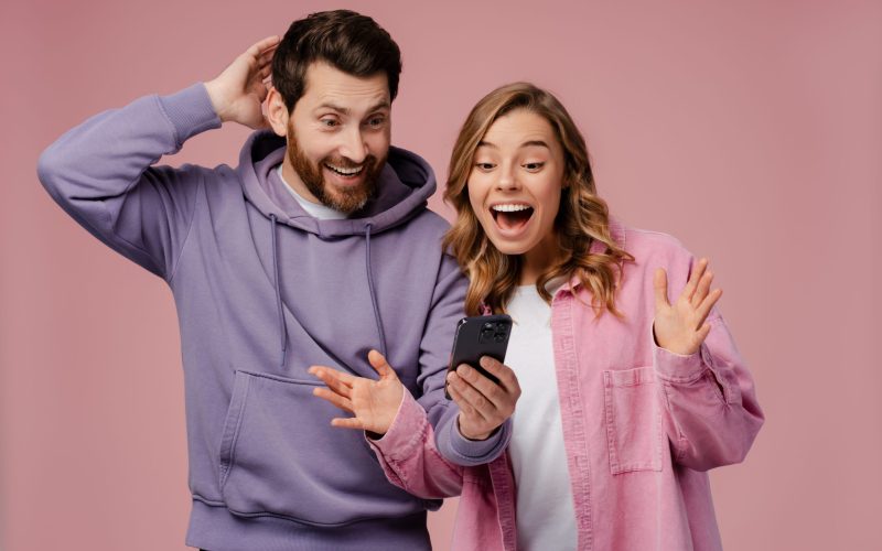 Portrait excited, smiling woman and man wearing stylish colorful clothes, holding mobile phone, using mobile app, win money standing isolated on pink background. Sport betting concept, online shopping
