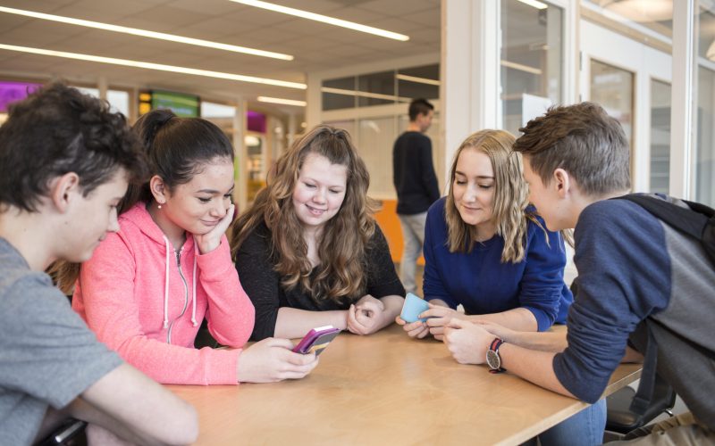 Group of teenagers engrossed in smartphones at a school table.