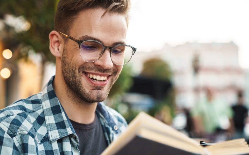 Smiling man enjoying free time and reading a book while sitting outdoors at coffee shop