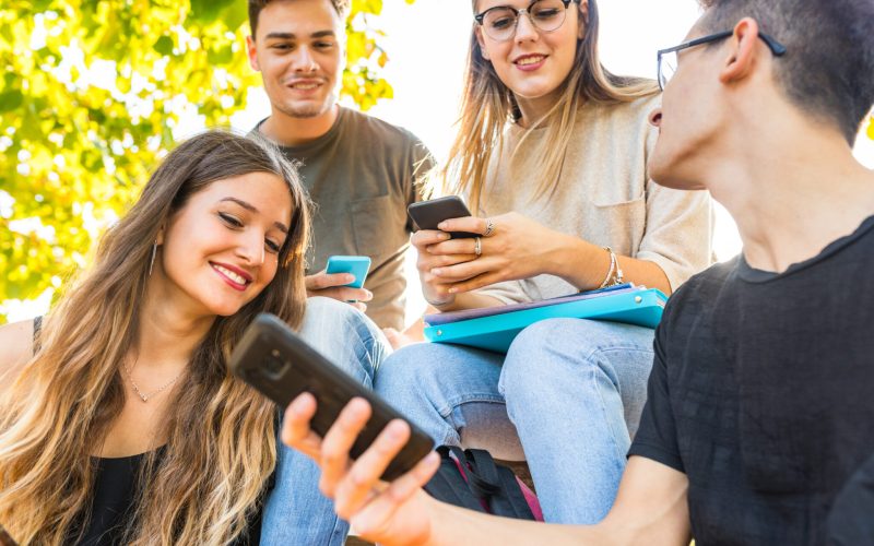 Teen group of friends with smartphones at park. Millennials best friends using mobile phones, addicted to technology and social media. Lifestyle and friendship concepts