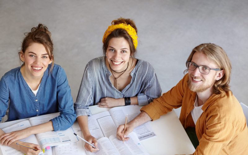 Two women and one bearded guy sitting together at table in front of opened books writing some notes with pencils making project together communicating with each other. Photo from above of three people